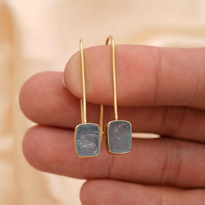 Puede incluir: Un par de pendientes colgantes dorados. Cada pendiente presenta una piedra preciosa rectangular de color azul claro engastada en un marco dorado. Los pendientes cuelgan de un fino alambre dorado curvado.