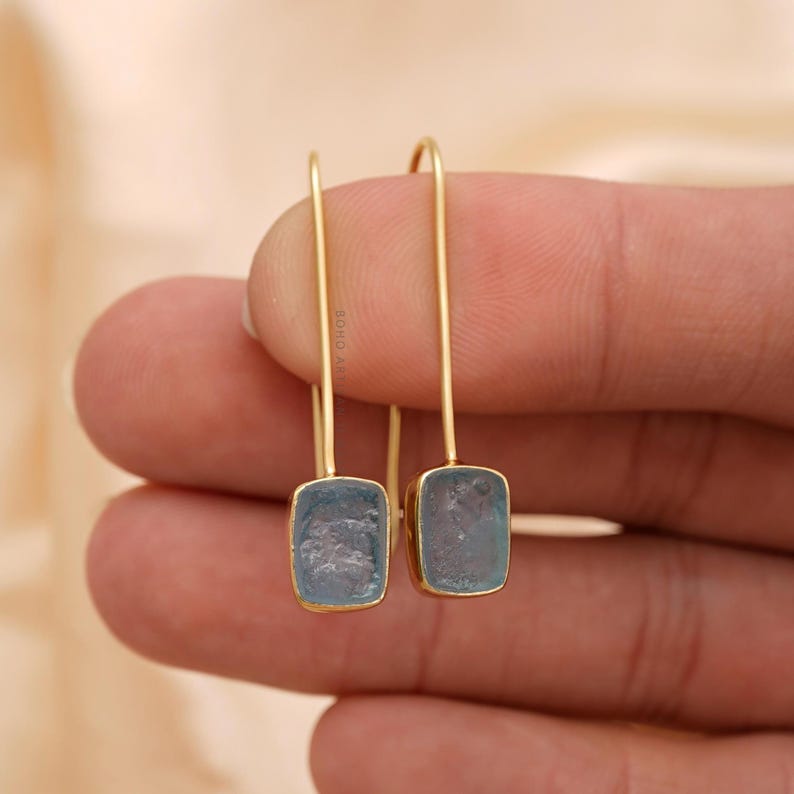 Puede incluir: Un par de pendientes colgantes dorados. Cada pendiente presenta una piedra preciosa rectangular de color azul claro engastada en un marco dorado. Los pendientes cuelgan de un fino alambre dorado curvado.