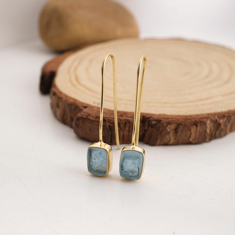 Puede incluir: Un par de pendientes colgantes dorados con piedras preciosas cuadradas de color azul claro. Los pendientes se exhiben sobre una superficie de madera con un fondo claro. Las piedras est&aacute;n engastadas en metal dorado.