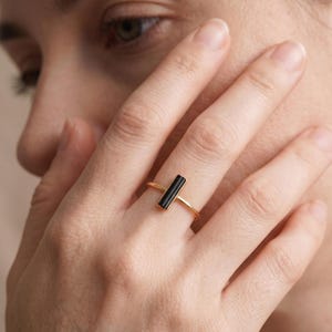 Pode incluir: Um anel de banda fina de ouro com uma única pedra preciosa preta retangular, orientada verticalmente e engastada no centro. A pedra tem uma superfície lisa e polida, mantida por um engaste de metal simples ligado à banda de ouro estreita.