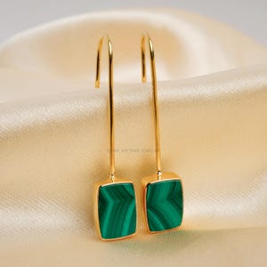 Puede incluir: Un par de pendientes colgantes dorados con piedras rectangulares de malaquita verde. Los pendientes tienen un dise&ntilde;o de gancho y se muestran sobre una tela satinada color crema. Joyas.