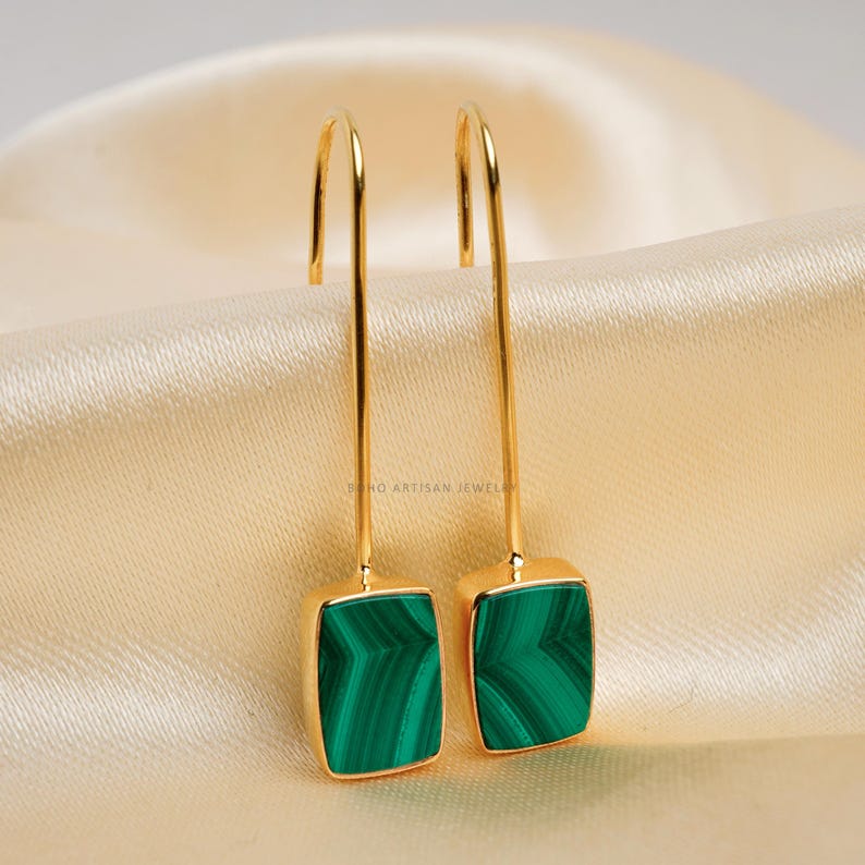 Puede incluir: Un par de pendientes colgantes dorados con piedras rectangulares de malaquita verde. Los pendientes tienen un dise&ntilde;o de gancho y se muestran sobre una tela satinada color crema. Joyas.