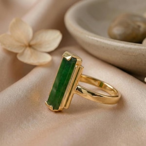 May include: A gold ring featuring a rectangular, emerald-green gemstone. The ring has a simple, elegant design with a polished gold band and setting. The gemstone is the focal point, showcasing its rich color and shape. The ring is displayed on a satin surface.