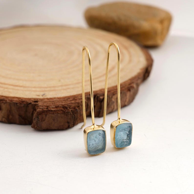 Puede incluir: Un par de pendientes colgantes dorados con gemas cuadradas de color azul claro. Los pendientes se exhiben sobre una superficie de madera, con un patr&oacute;n de vetas naturales. Las gemas tienen una apariencia ligeramente texturizada.