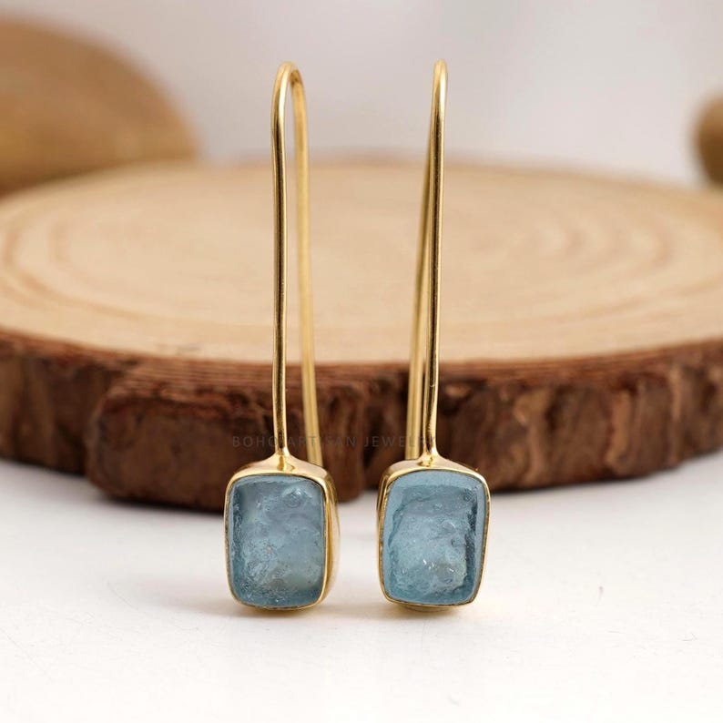 Puede incluir: Un par de pendientes colgantes dorados con piedras preciosas rectangulares de color azul claro. Los pendientes tienen un dise&ntilde;o sencillo y elegante, con un gancho largo y curvado y un engaste de bisel para las piedras preciosas.