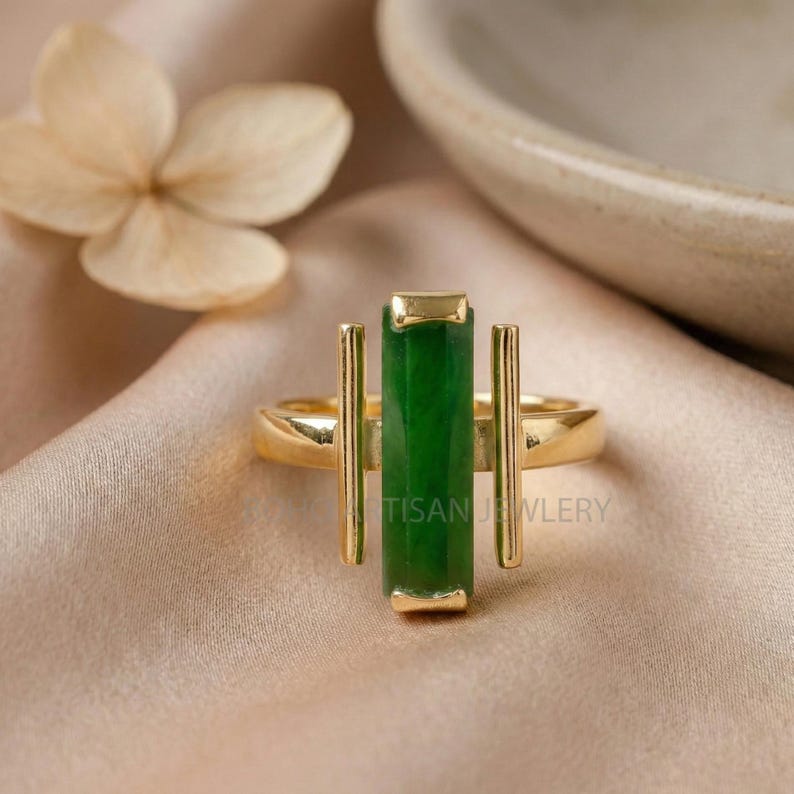 May include: A gold ring featuring a rectangular, vibrant green gemstone. Two vertical gold bars flank the green stone, with the text "TISAN JEWLERY" visible. The ring is set against a soft, neutral background.