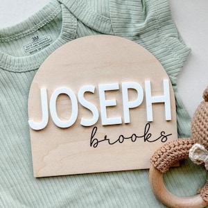 May include: A wooden sign with the name "JOSEPH" in white block letters and the last name "brooks" in cursive. A brown crochet bear rattle and a light green baby onesie are also in the image.