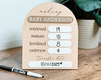 May include: An arched wooden sign with the text "making BABY ANDERSON" and rows labeled "retrieved 19", "mature 10", "fertilized 8", "embryos 4", and "Transfer date 10/11/2025" with a heart icon, accompanied by a black dry-erase marker.