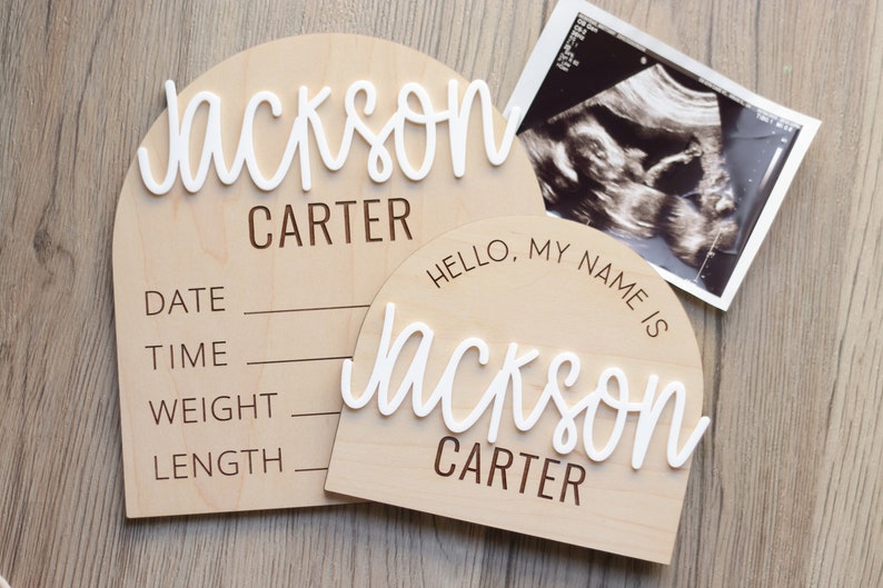 May include: Two arched light wood signs with white 3D script lettering and dark brown engraved text. The larger sign features the name Jackson Carter with fields for date, time, weight, and length. The smaller sign reads Hello, My Name Is Jackson Carter.