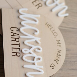 May include: Two arched wooden name plaques featuring the laser-engraved text HELLO, MY NAME IS and CARTER. Each plaque displays a raised, white, 3D-style script name reading Jackson, layered over the light-colored wood surface.