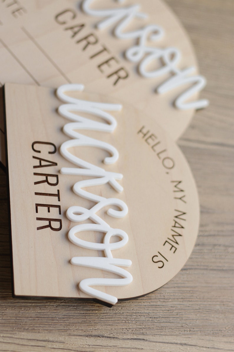 May include: Two arched wooden name plaques featuring the laser-engraved text HELLO, MY NAME IS and CARTER. Each plaque displays a raised, white, 3D-style script name reading Jackson, layered over the light-colored wood surface.