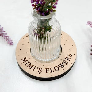 May include: A wooden coaster with the text "Mimi's Flowers" engraved on it. A small glass vase with purple flowers sits on top of the coaster.
