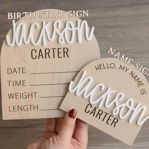 May include: Two arched wooden signs with light wood grain. The larger sign features the text BIRTH STAT SIGN, Jackson in white 3D script, Carter, and lines for Date, Time, Weight, and Length. The smaller sign features NAME SIGN, Hello, My Name Is, Jackson in white 3D script, and Carter.