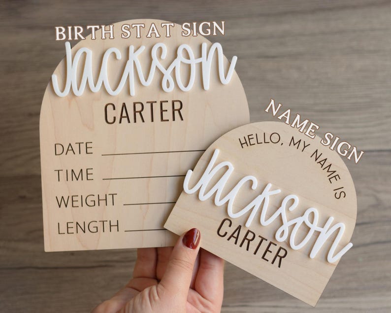 May include: Two arched wooden signs with light wood grain. The larger sign features the text BIRTH STAT SIGN, Jackson in white 3D script, Carter, and lines for Date, Time, Weight, and Length. The smaller sign features NAME SIGN, Hello, My Name Is, Jackson in white 3D script, and Carter.