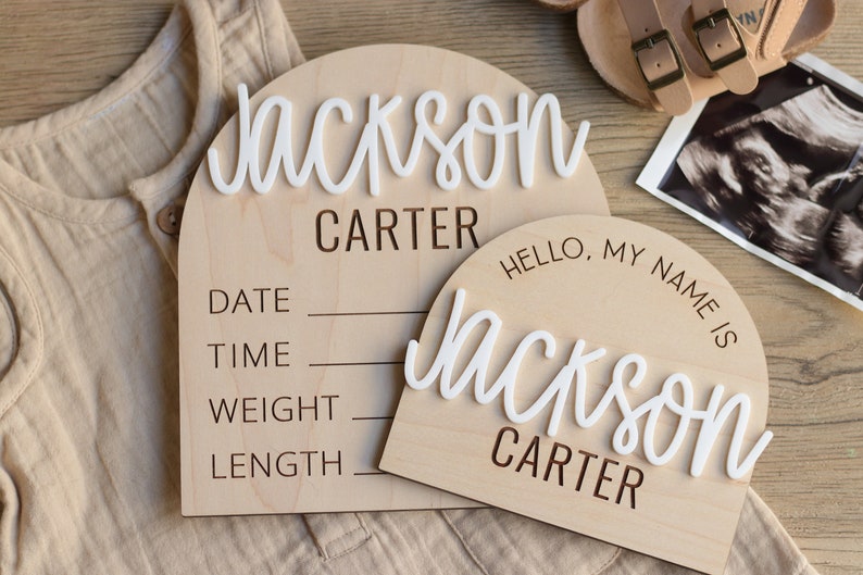 May include: Two arched wooden birth announcement signs with light wood grain. The larger sign features the white 3D name Jackson, the printed name CARTER, and lines for DATE, TIME, WEIGHT, and LENGTH. The smaller sign says HELLO, MY NAME IS.