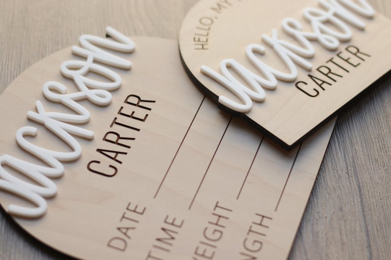 May include: Two circular wooden birth announcement signs featuring the name Jackson Carter in raised white script lettering. The signs include engraved text that reads Hello, My Name Is, Date, Time, Weight, and Length with lines for writing.