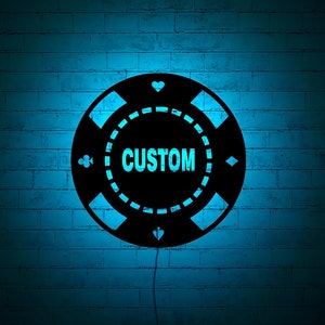 Op de afbeelding: Zwarte pokerchip met een witte rand en het woord "CUSTOM" in witte letters. De chip bevindt zich op een blauwe bakstenen muur.