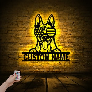 May include: A black metal silhouette of a German Shepherd dog wearing American flag sunglasses. The dog is peeking over a metal sign that says "CUSTOM NAME". The sign is illuminated with warm yellow light.