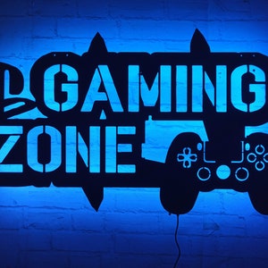 Könnte beinhalten: Schwarzes Metallschild mit den Worten "Gaming Zone" und einer Silhouette eines Videospiel-Controllers. Das Schild ist mit blauem Licht beleuchtet.