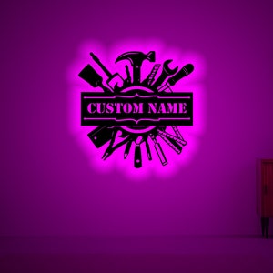 May include: Black metal wall art featuring a circle of tools with a banner that says "CUSTOM NAME". The art is illuminated with a pink glow.