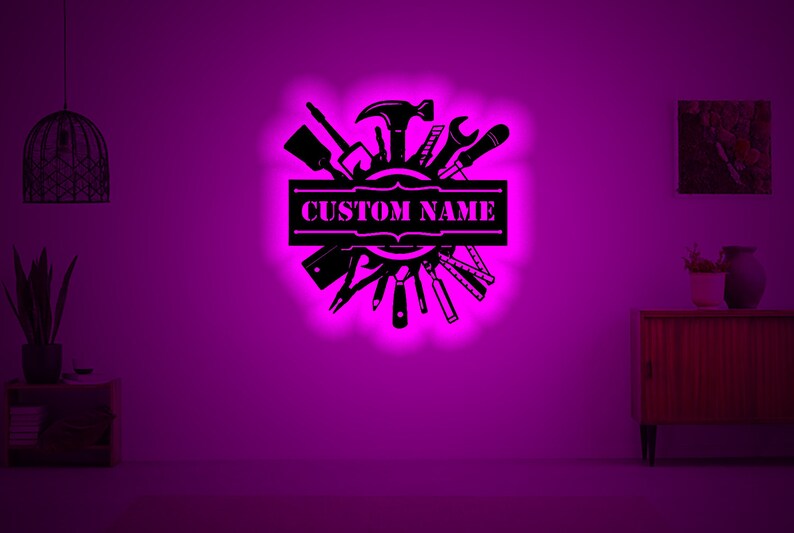 May include: Black metal wall art featuring a circle of tools with a banner that says "CUSTOM NAME". The art is illuminated with a pink glow.