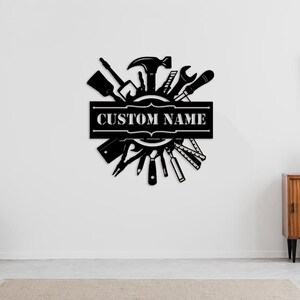 May include: Black metal wall art featuring a circle of tools, including a hammer, wrench, screwdriver, and saw. The circle is surrounded by a banner with the text "CUSTOM NAME".
