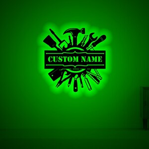 May include: A black metal wall decor with a circle of tools and a banner that says "CUSTOM NAME". The tools include a hammer, wrench, screwdriver, and saw. The background is a green wall.
