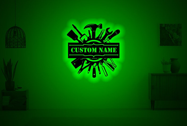 May include: A black metal wall decor with a circle of tools and a banner that says "CUSTOM NAME". The tools include a hammer, wrench, screwdriver, and saw. The background is a green wall.