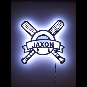 May include: A white illuminated baseball themed sign with two crossed bats and a baseball in the center. The name "Jaxon" is written on a banner above the baseball.
