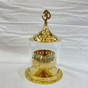 May include: A gold metal incense burner with a glass dome and an Om symbol on top. The burner has a decorative design with intricate details.