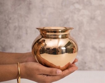 Può includere: Un vaso di metallo lucido color oro, con un corpo arrotondato e un bordo svasato. Il vaso è tenuto in mani a coppa e la superficie riflette l'ambiente circostante. Un braccialetto d'oro adorna il polso.