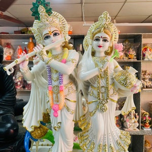 May include: White marble statue of Radha Krishna, a Hindu deity couple. The statue depicts Radha holding a lotus flower and Krishna playing a flute. The statue is decorated with gold accents and colorful details.