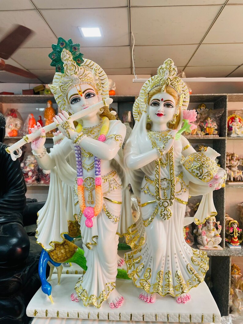 May include: White marble statue of Radha Krishna, a Hindu deity couple. The statue depicts Radha holding a lotus flower and Krishna playing a flute. The statue is decorated with gold accents and colorful details.