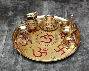 May include: A golden brass puja thali set with various religious items. The thali features red and green accents with the Om symbol and other religious symbols. The set includes a small bell, a lamp, and other decorative items.