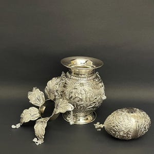 May include: A silver-colored, ornate metal vessel with a wide, open top, intricate carvings, and a matching egg-shaped container. The vessel has a floral-like attachment and is set against a dark background. The design features detailed patterns and hanging chains.