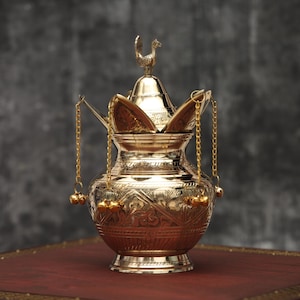 May include: A gold-colored incense burner with intricate floral patterns and a rooster-shaped finial. The burner has a pot-like shape with a lid and hanging chains with bells. It sits on a dark red surface against a gray background.