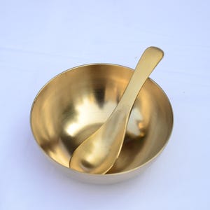 Op de afbeelding: Een gouden messing kom met een bijpassende lepel erin. De kom is rond en heeft een glad, gepolijst oppervlak. De lepel is gebogen en heeft een vergelijkbare afwerking. De items staan tegen een witte achtergrond.