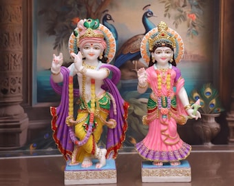 May include: A pair of intricately detailed, hand-painted figurines depicting Krishna and Radha. Krishna is playing a flute and Radha is holding a lotus flower. Both figurines are standing on a white base with gold accents.