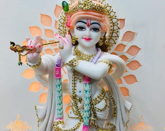 May include: A white marble statue of Krishna playing a flute. He is adorned with colorful jewelry and a green feathered headdress. The statue is set against a white background with a decorative pattern.