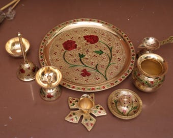 Puede incluir: Un juego de thali de metal dorado con un diseño decorativo de flores rojas y hojas verdes. El juego incluye un plato grande, un tazón pequeño, un diya, una cuchara y una campana.