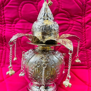 May include: A silver decorative pot with intricate floral designs and a bird finial. The pot has a round body and a wide base. It is adorned with dangling chains and bells.