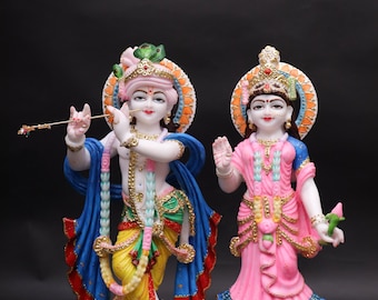 May include: Two ornate statues of Hindu deities. One figure, in yellow and blue garments, holds a flute. The other, in pink and blue, holds a flower. Both statues have halos and are on white pedestals.