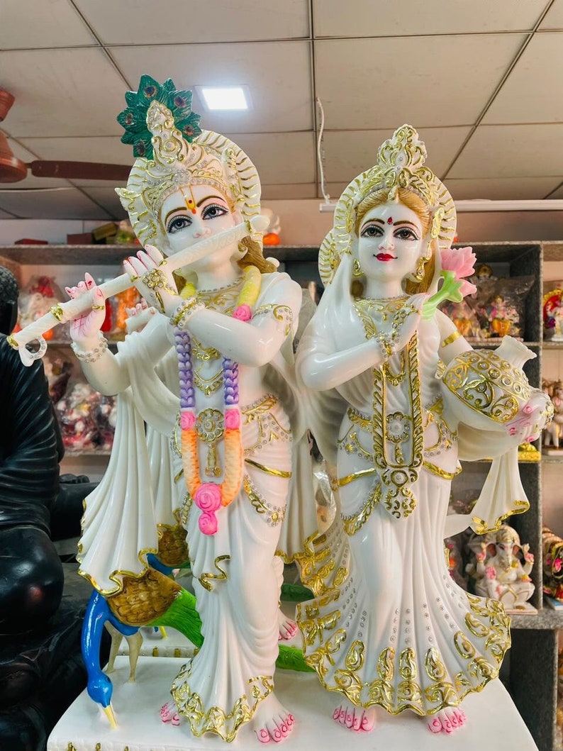 May include: White marble statue of Radha Krishna, a Hindu deity couple. The statue depicts Radha holding a lotus flower and Krishna playing a flute. The statue is decorated with gold accents and intricate details.