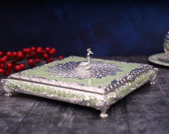 Può includere: Una scatola decorativa argentata e verde con un motivo a pavone sul coperchio. La scatola ha quattro piedini ed è realizzata in metallo.