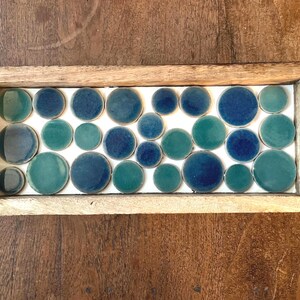 May include: A rectangular wooden tray filled with round, glazed ceramic tiles. The tiles are in shades of teal and blue, arranged in a grid pattern. The tray's frame is a light, natural wood color.