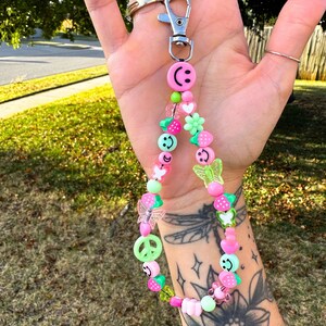 May include: A beaded keychain with a silver clasp. The keychain features a variety of colorful beads, including smiley faces, strawberries, butterflies, and flowers in shades of pink, green, and clear.
