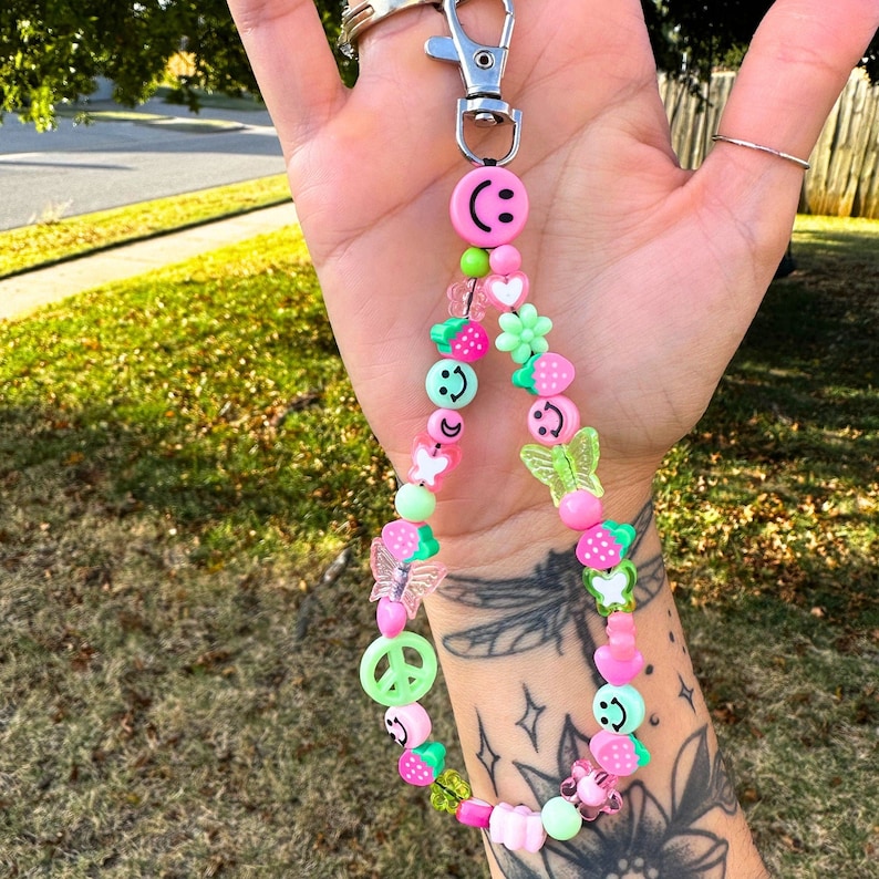 May include: A beaded keychain with a silver clasp. The keychain features a variety of colorful beads, including smiley faces, strawberries, butterflies, and flowers in shades of pink, green, and clear.