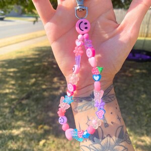 May include: A pink beaded keychain with a silver clasp. The keychain features various beads, including smiley faces, hearts, flowers, butterflies, and fruit slices. The color palette is primarily pink, with accents of blue and purple.