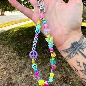 May include: A colorful beaded keychain with a silver clasp. The keychain features various beads, including smiley faces, flowers, butterflies, and the word "ONYX". The design incorporates a peace sign and fruit-shaped beads.