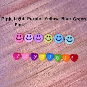 May include: A collection of colorful smiley face and heart-shaped embellishments. The smiley faces are pink, light purple, purple, yellow, blue, and green. The hearts are pink, light pink, purple, yellow, blue, green, orange, and red. The text above the embellishments reads "Pink Light Purple Yellow Blue Green Pink".